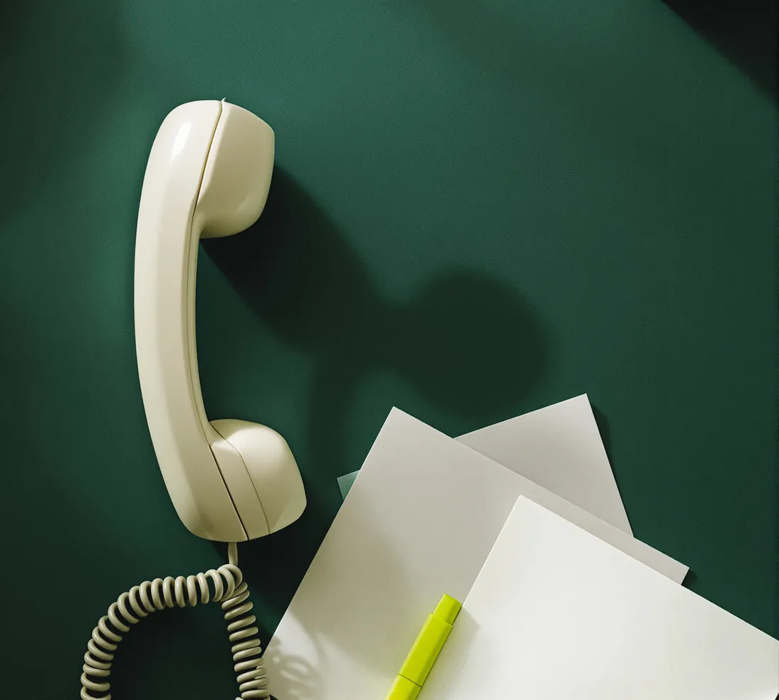 A cream telephone handset lying off the hook on a dark green desk, next to loose sheets of paper and a lime highlighter.